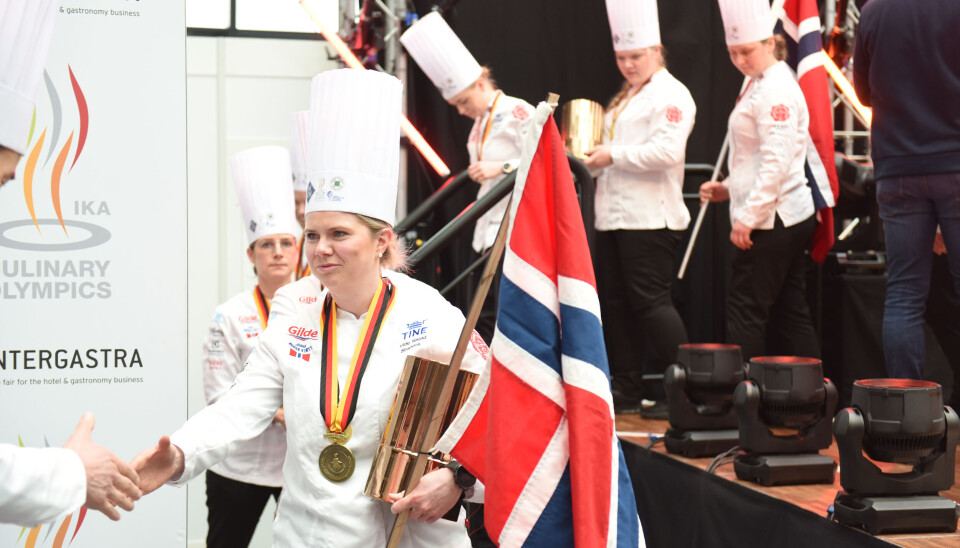 Det var en tydelig rørt Vilde Nistad Strandos som tok imot bronsemedalje for sin innsats på De Norske Kokkelandslagene Community Catering under Kokke-OL 2024 i Stuttgart. Jente med kokkejakke og kokkehatt med madalje rundt halsen og pokal og flagg i hånda.