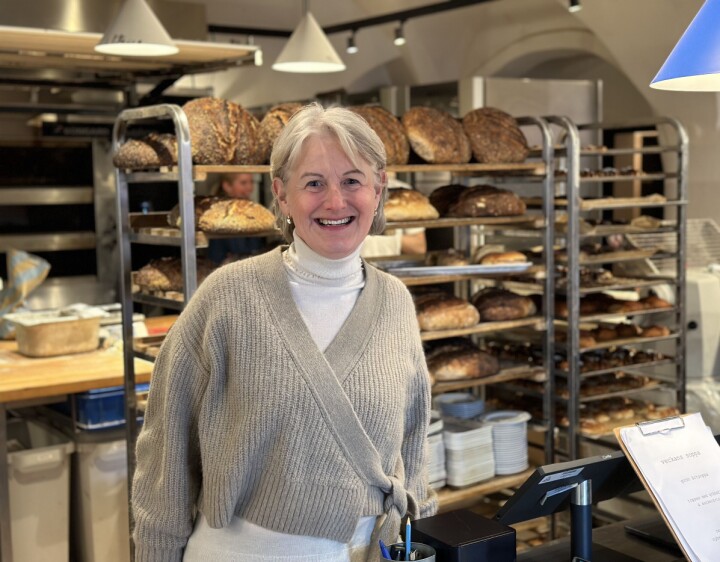 Kjedekoordinator i Din Baker, Ida Ribler, inviterer til butikksamling. Dame med grå genser i et bakeri med stekte brød på traller i bakgrunnen.
