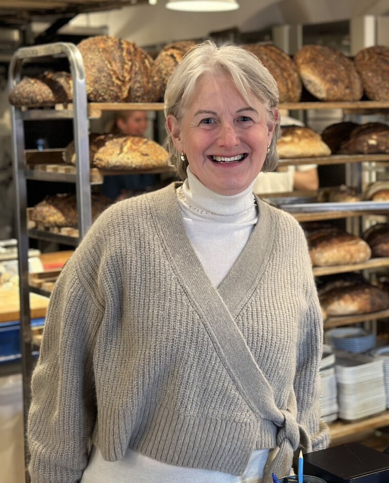 Ida Ribler har to måneder bak seg som kjedekoordinator i Din Baker. Hun tar av seg hatten for de som jobber i bransjen. Dame med grå genser i et bakeri med stekte brød på traller i bakgrunnen.