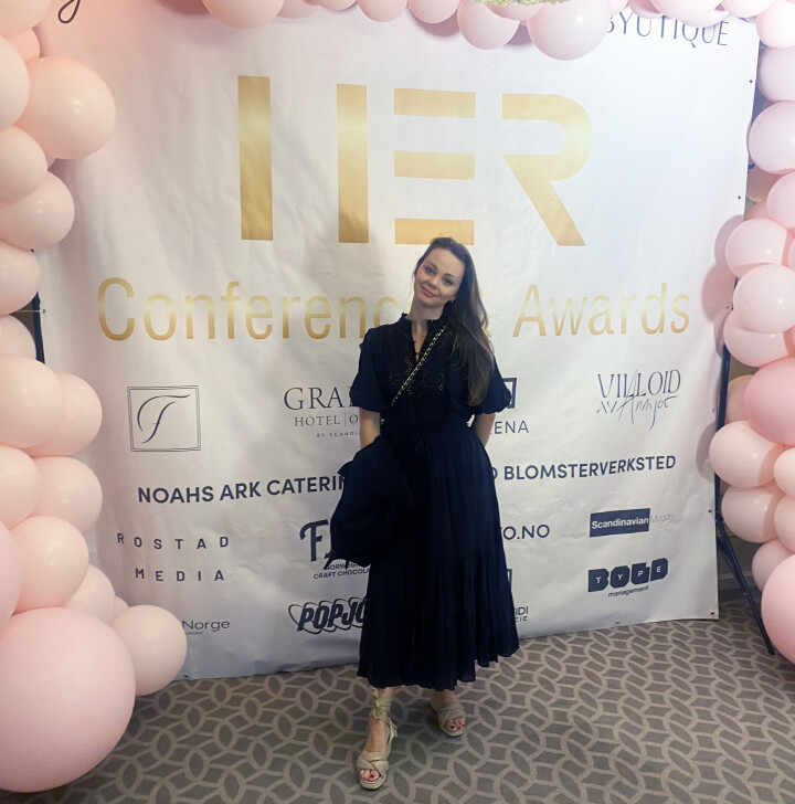Eier og daglig leder av Aanerud Bakeri, Hanna Marie Halvorsen Bergan, deltok på Her Conference & Awards i Oslo mandag 13. mai. Dame i snart kjole foran sponsorvegg med rosa ballonger rundt.