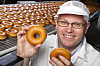 Mann med briller som holder to donuts foran en produksjonslinje full av donuts.
