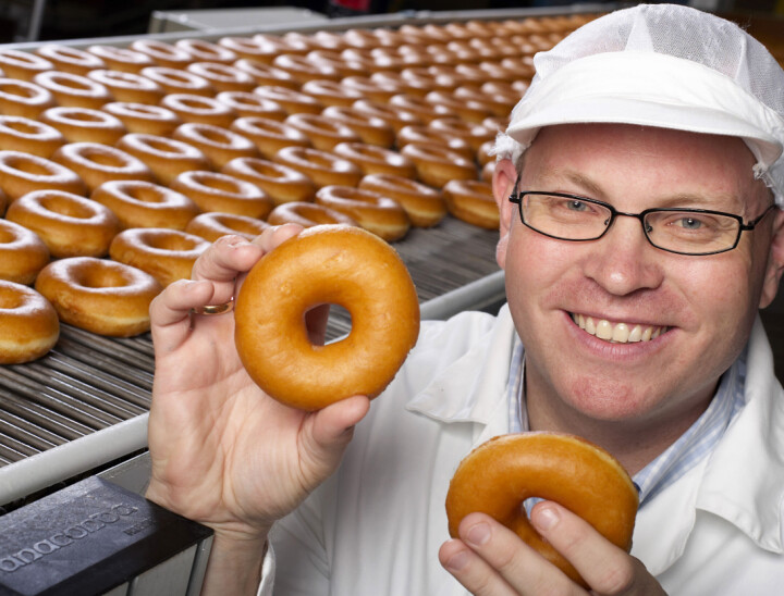 Bernt Ove Søvik og Millba øker staben. Mann med briller som holder to donuts foran en produksjonslinje full av donuts.