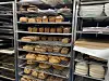 Flere hyller fulle av nystekte brød og bakervarer på metallstativ i et bakeri.