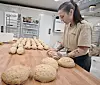 Baker former brøddeig på trebord foran rekker av deigemner i bakeri