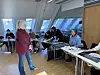 Instruktør snakker til voksne deltakere som sitter ved pulter i et lyst klasserom.