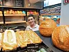 Ansatt bak disk i bakeri med store brød og bakervarer utstilt i forgrunnen.