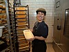Smilende baker i arbeidsklær holder en form med brøddeig foran et stativ med brett i et bakeri.