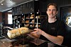 Baker holder et fat med store kakestykker foran brødhylle i bakeri
