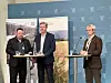 Tre menn ved podier under en pressekonferanse foran blå vegg med riksløve-symboler.