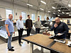 Fire menn i en verkstedhall ser på en arbeidsbenk med materialer og dokumenter.