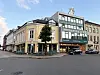 Bygate med historiske bygninger, en moderne hjørnefasade og parkerte biler under klar himmel.