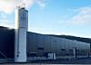 Grå industribygning med hvit silo ved siden av, sett utendørs under blå himmel.