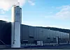 Grå industribygning med hvit silo ved siden av, sett utendørs under blå himmel.