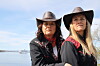 To kvinner i svarte klær og cowboyhatter står ved sjøen med en kystlinje bak.