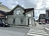 Grønt hjørnehus i tre ved en gate med bil, fortau og moderne bygninger i bakgrunnen.