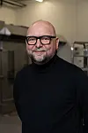 Mann med briller og svart høyhalset genser står foran maskiner i et industrielt lokale.