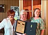 Tre kvinner poserer innendørs, en holder en innrammet utmerkelse og en medalje fra G. Johnsen.