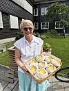 Smilende kvinne holder en kurv med boller i en hage foran et hus.