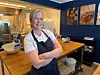 En kokk i forkle står smilende i et restaurantkjøkken med arbeidsbenk og kjøkkenutstyr.