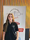 Kvinne står foran et banner med Baker & Konditori- og Idun Industri-logoer i et rom med trelameller.