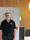 Mann i svart skjorte står foran et banner med bakeri- og konditorlogo i et innendørs lokale.