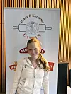 Ung kvinne står foran et banner med «Baker & Konditori Campus» i et rom med vertikale trepaneler.
