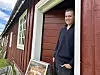 Mann i mørk genser står i døråpningen til et rødmalt hus med hvite vinduer.