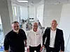 Tre menn står i et lyst kontorlandskap med glassvegger og skrivebord i bakgrunnen.
