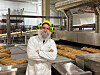 En bakerimedarbeider i hvit uniform og verneutstyr står ved brett med brød i en industriell bakerihall.
