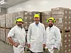 Tre ansatte i verneutstyr står i en produksjonshall med stablede paller i bakgrunnen.