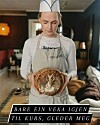Person i bakerlue og forkle holder et dekorert brød i et innendørs kjøkkenmiljø.