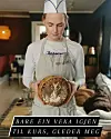 Person i bakerlue og forkle holder et dekorert brød i et innendørs kjøkkenmiljø.