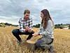 To personer kneler i et kornåker og ser på avling i en kurv.