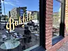 Gullfarget skilt i et butikkvindu på en rød teglbygning, med speilinger av en bygate.