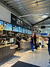 Kunder ved ferskvaredisker i et lyst innendørs matmarked med digitale skjermer.
