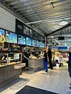 Kunder ved ferskvaredisker i et lyst innendørs matmarked med digitale skjermer.