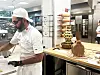 Baker arbeider ved en benk i et profesjonelt kjøkken med brødskulptur i bakgrunnen.