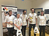 Seks bakerei hvite uniformer poserer med medaljer og premier i et rom.