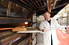 Baker i hvitt holder en trespade med et brød foran en varm steinovn.
