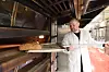 Baker i hvitt holder en trespade med et brød foran en varm steinovn.