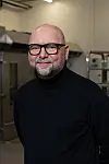 Smilende mann med briller og svart genser foran rustfritt kjøkkenutstyr.