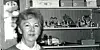 Svart-hvitt portrett av en kvinne foran en hylle med pokaler og glass.