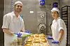 To bakeriansatte står bak et brett med ferske bakverk i et industrikjøkken.