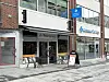 Gatefasade med kafé, utebord, butikkvinduer og skilt langs en urban gågate.