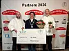 Tre menn poserer med blomster, pokal og stor sjekk foran en sponsorvegg.