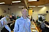 Smilende mann i blå skjorte foran deltakere i et lyst konferanselokale.