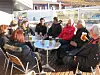 Mennesker rundt et kafébord på en utendørs terrasse med drikke og bymiljø i bakgrunnen.