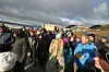 Folkemengde i ytterklær samlet foran fjell og bygninger under en skyfull himmel.