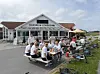 Gruppe mennesker ved picnicbord utenfor en kafébygning med sykler og parasoller.