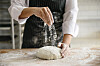 Baker drysser mel over en surdeig på en melet arbeidsbenk.